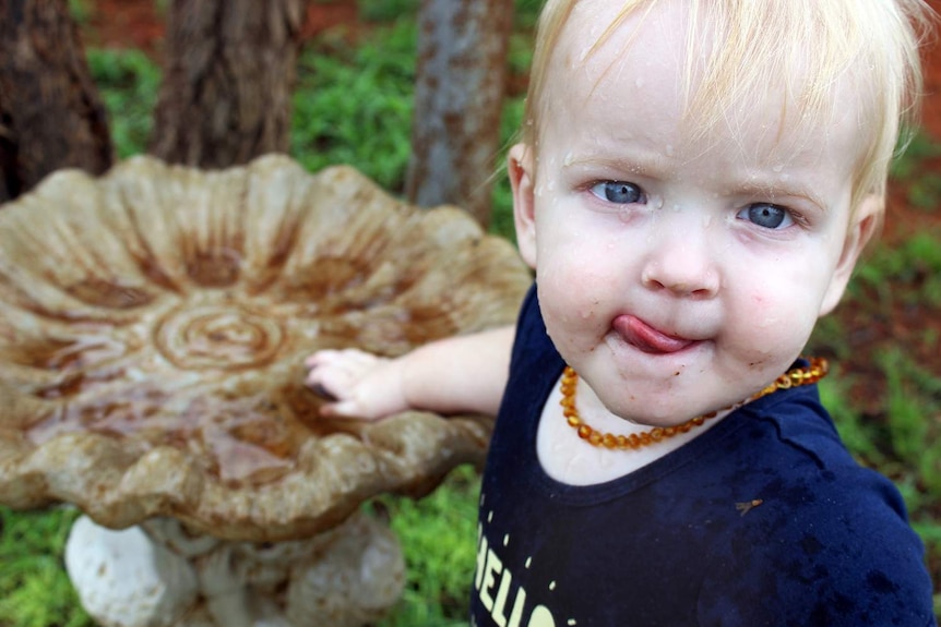 Toddler Toby Campbell looks up with raindrops on his face and licks his lips as his hand plays in water in a birdbath.