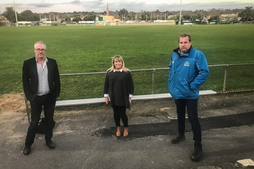 A middle-aged man, a short blonde woman and another taller man of middle age standing in front of a sports field.