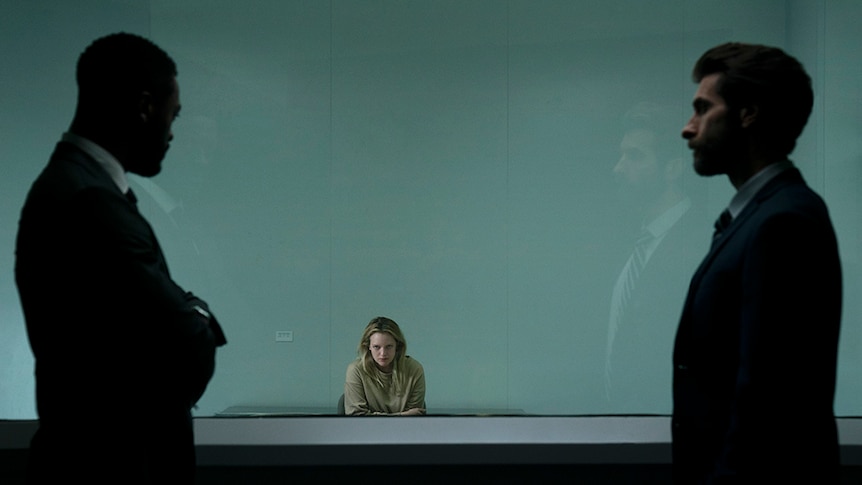 Two silhouetted men in suits stand behind glass window and observe a serious looking seated woman in interrogation room.