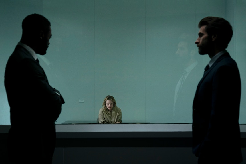 Two silhouetted men in suits stand behind glass window and observe a serious looking seated woman in interrogation room.