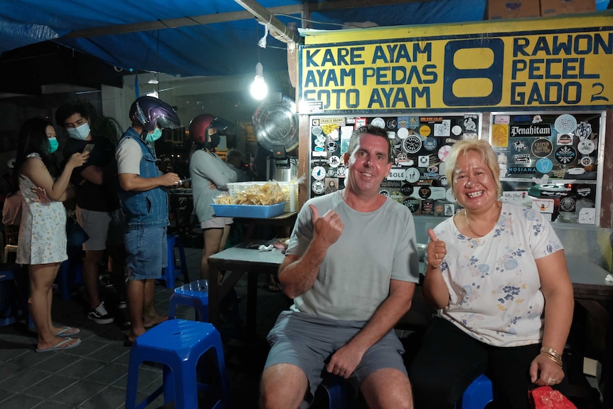 A man and a woman give a thumbs up in front of a food stall.