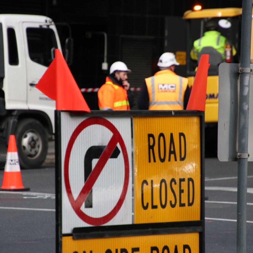 A road closed sign with two construction workers in the background.