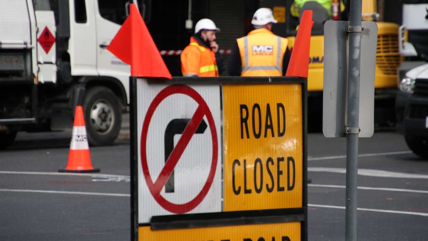A road closed sign with two construction workers in the background.