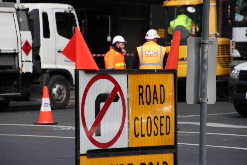 A road closed sign with two construction workers in the background.