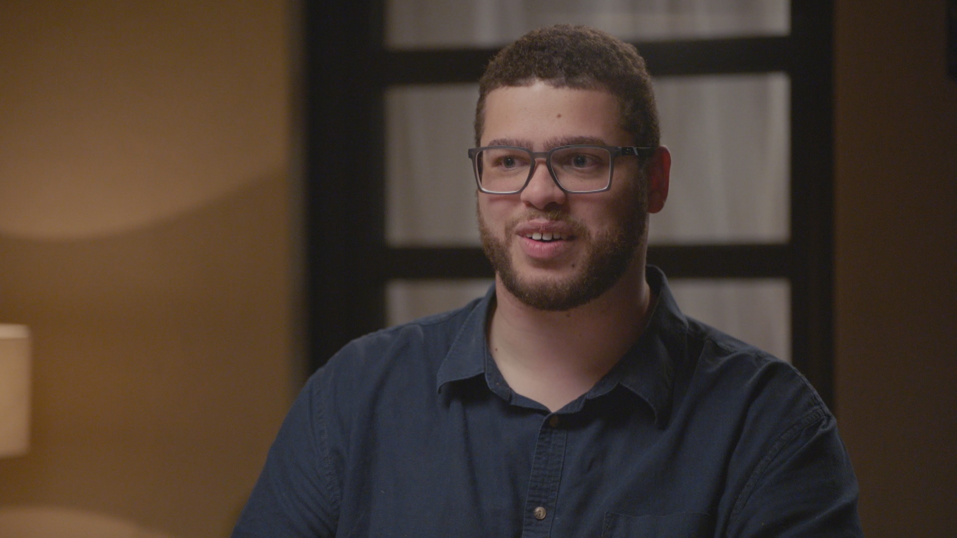 Jordon Steele-John wearing a navy collared shirt looking to the left of the camera, a lamp and door behind him are out of focus
