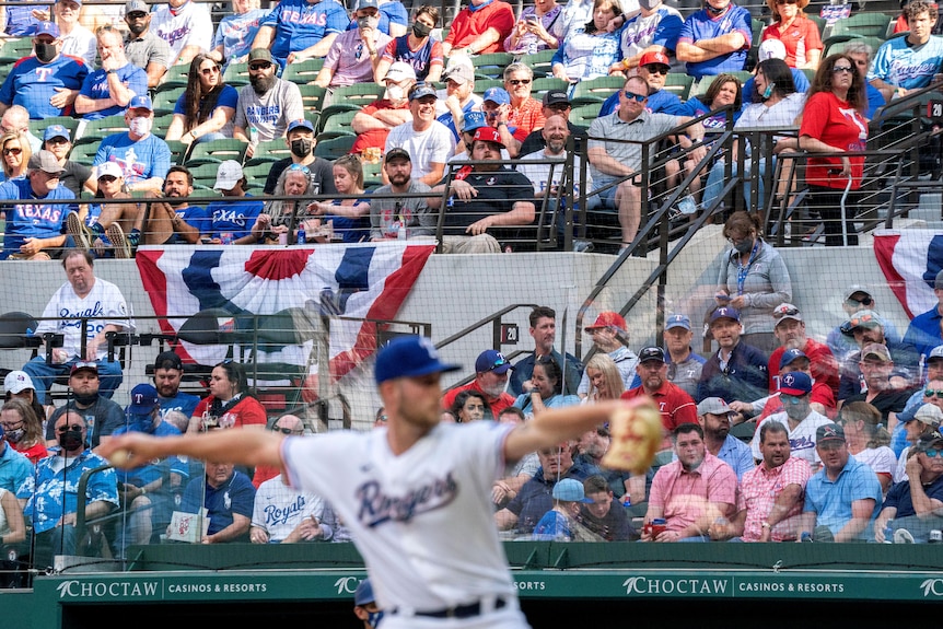 Un hombre vestido de blanco lanza la pelota con los fanáticos mirando en el fondo