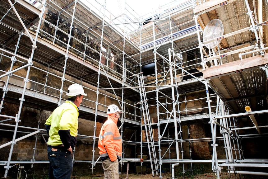 Two builders survey a courtyard covered in scaffolding.