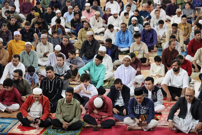 A group of people sitting on mats, some of them praying, inside an indoor sports stadium.
