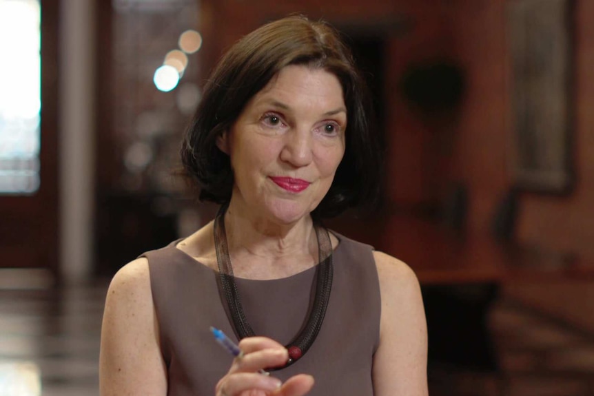 MCU of Michele Levine, wearing a sleeveless brown dress, sitting in a meeting room