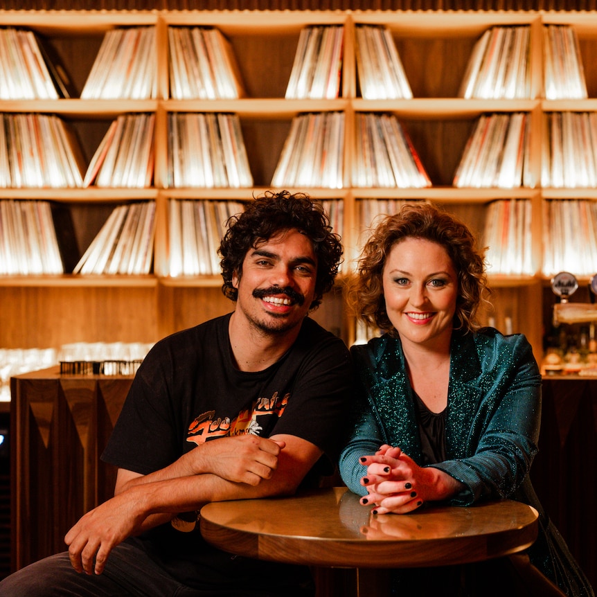 Tony and Zan sit at a table in a bar with vinyl records on the wall