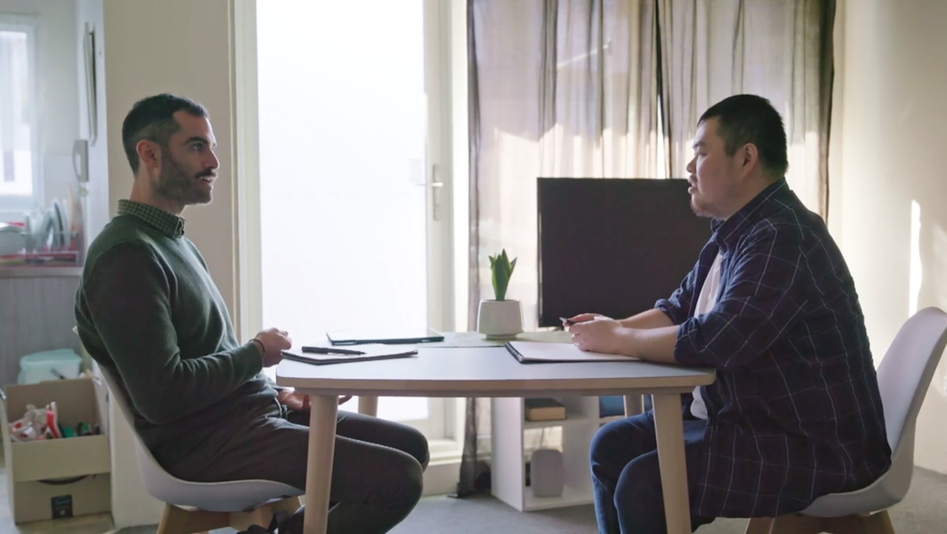 Two men sit on either side of a table in a small kitchen and talk, and notepad and pen in between them. 