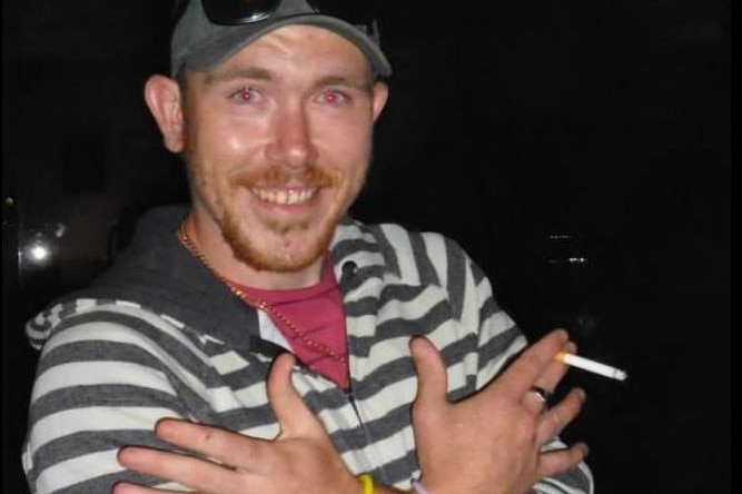 Andrew smiles into the camera, holding a lit cigarette and crossing his arms.