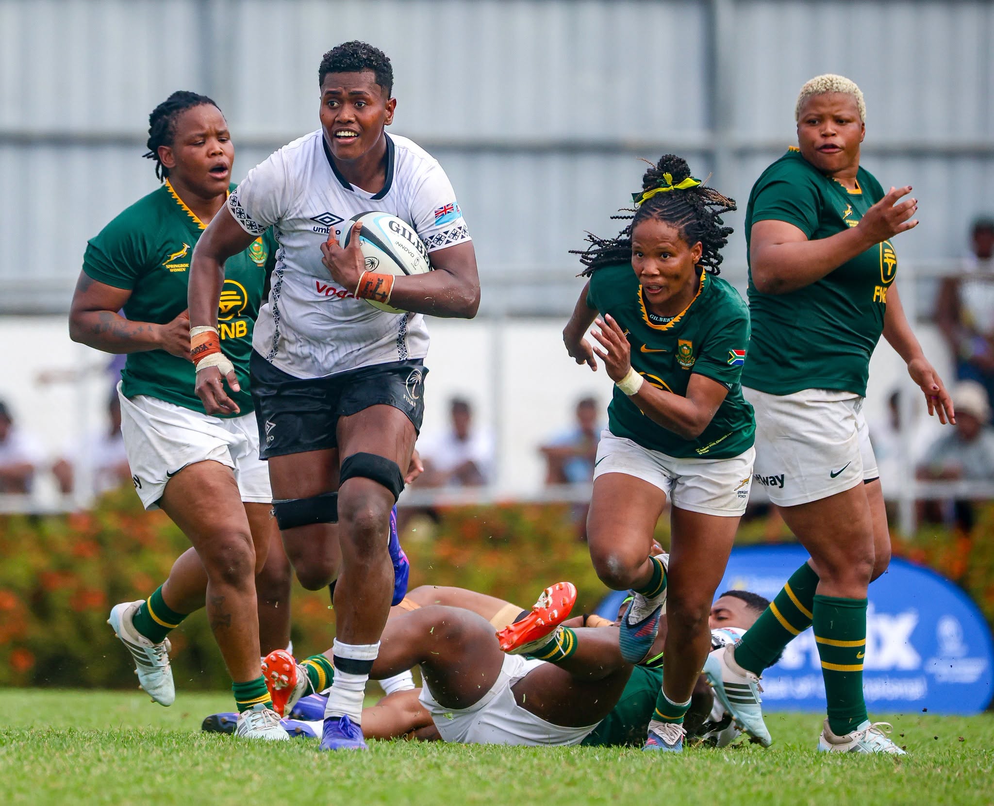A Fijian woman wearing a white rugby uniform holding a rugby ball runs on a field while a woman in green uniform chases her