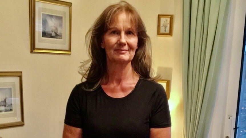 An older woman with long brown hair standing near a wall of photos.