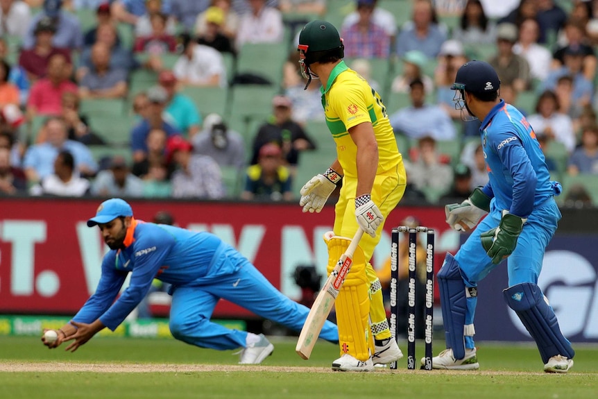 An Indian fielder takes a catch low at slip as the Australian batsman and the wicketkeeper look on.