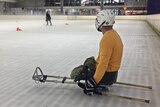 A para ice hockey player strapped into a black plastic sled watches his hockey coach demonstrate a drill on the ice.