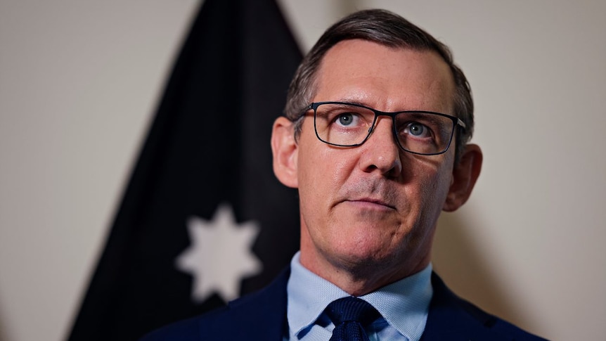 A middle-aged man in a navy suit and tie standing in front of a flag with his head tilted. It's a head-and-shoulders shot.