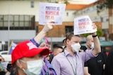 A crowd with masks on stands outside a hospital, some with signs held above their heads.