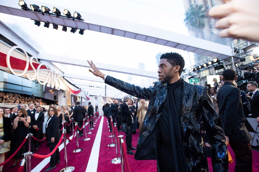 Chadwick Boseman waves to fans on the red carpet. The fans are behind a partition, with Boseman separated by velvet ropes.