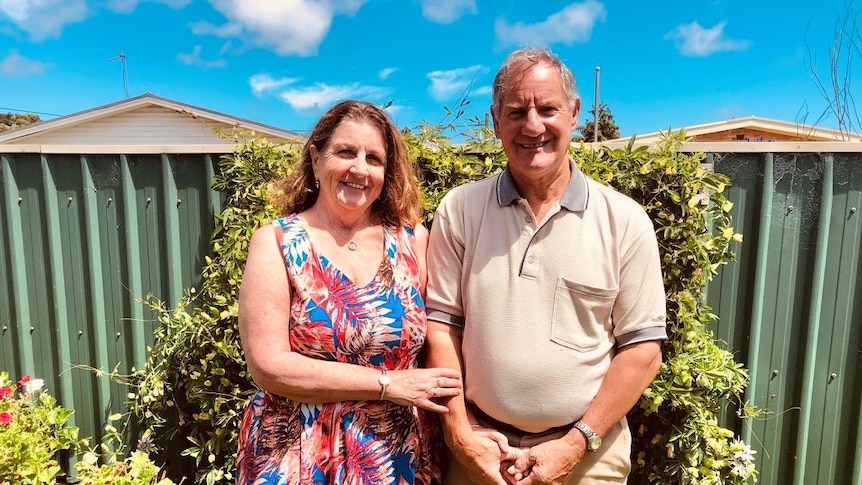 Liz and Ray stand in their backyard, hands held tightly in front. Both are smiling and look happy. There is a bright blue sky.