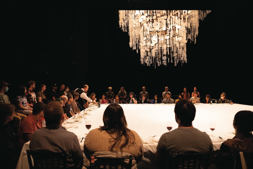 Rows of guests sit ten-per-side at a giant table lined with a white tablecloth and place settings in a darkened theatre.
