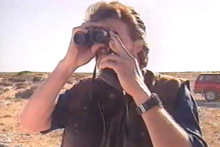 A man stands on what appears to be the top of a cliff, looking through a pair of binoculars.