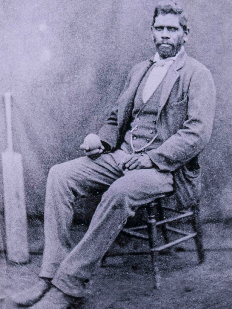 A seated Unaarrimin aka Johnny Mullagh wearing a suit with a cricket bat in the background.
