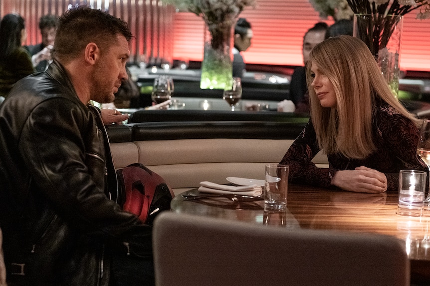 Man with dark hair and black leather jacket sits opposite woman with long blonde hair and black patterned shirt at dining booth.