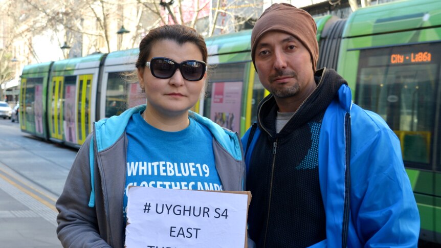 A Uyghur couple wearing bright blue jackets stand in front of a green Melbourne tram.