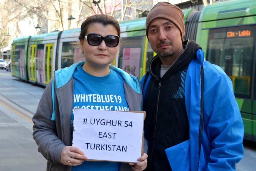 A Uyghur couple wearing bright blue jackets stand in front of a green Melbourne tram.