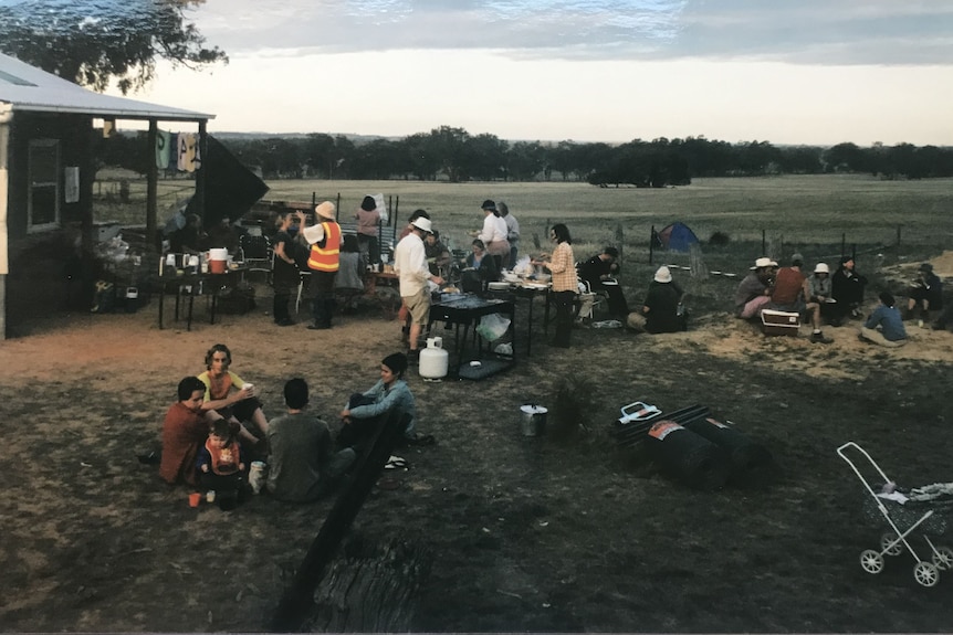 About 25 people sit in groups eating and chatting at dusk in the bare paddock