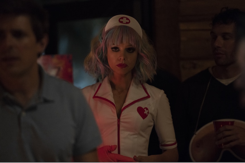 Dark interior shot showing young woman with pastel-streaked blonde bob, red lips, dark eyes, wearing Halloween nurse costume.