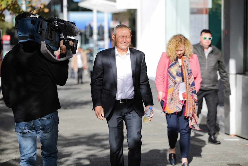 A man in a blazer and white shirt smiles as he's being filmed by a TV camera.