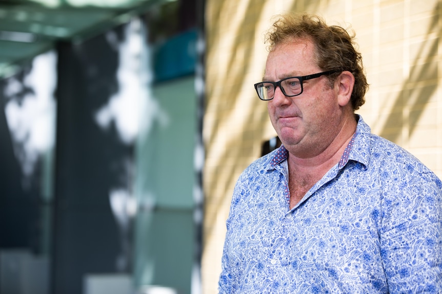 A man in a blue shirt and glasses looks to the left with a furrowed expression.