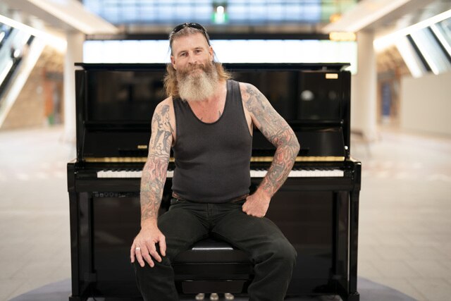 A man with a beard standing up in front of a piano in a train station.