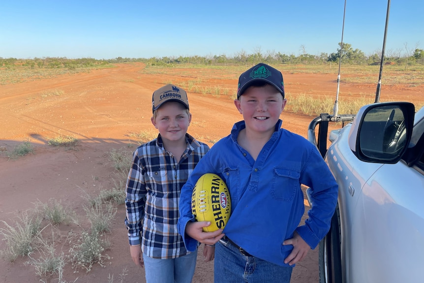 Two young boys wearing caps, one holding a football, standing next to a car on a dirt track