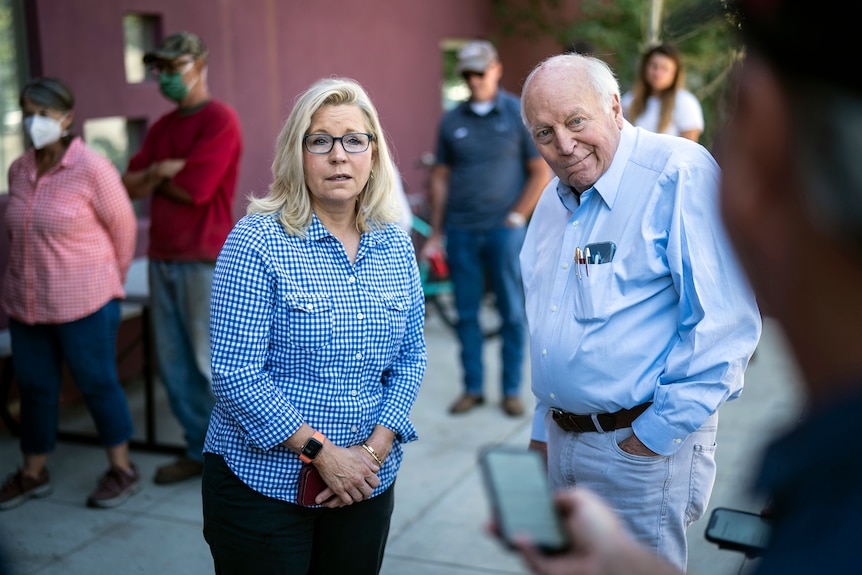 A middle aged woman and an elderly man stand next to each other near a queue of voters, some in face masks.