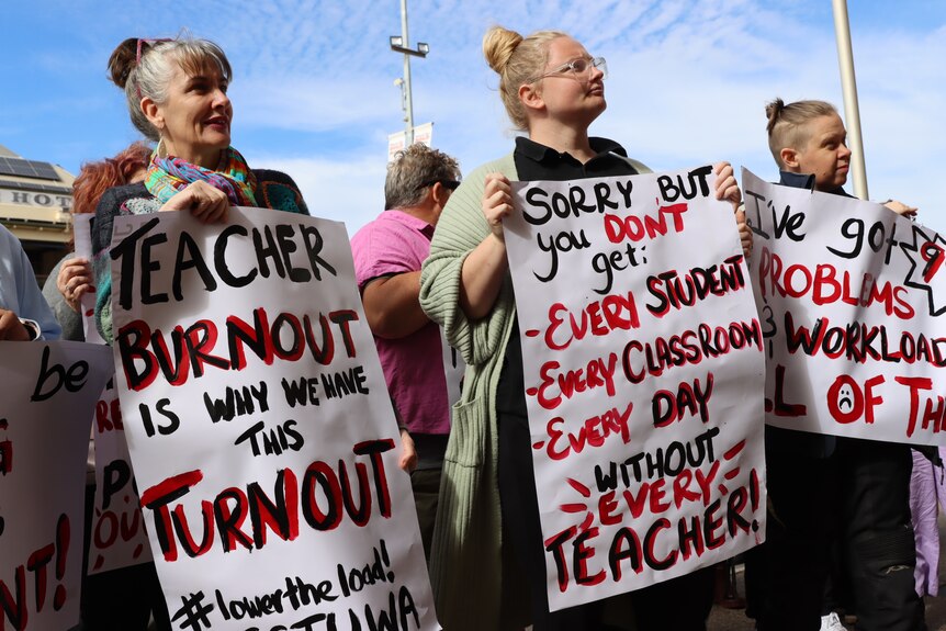Three people hold signs about teacher burnout at a rally for better pay and conditions. 