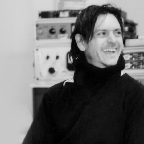 black and white photo of a smiling man in a black jumper, extensive sound studio equipment behind him