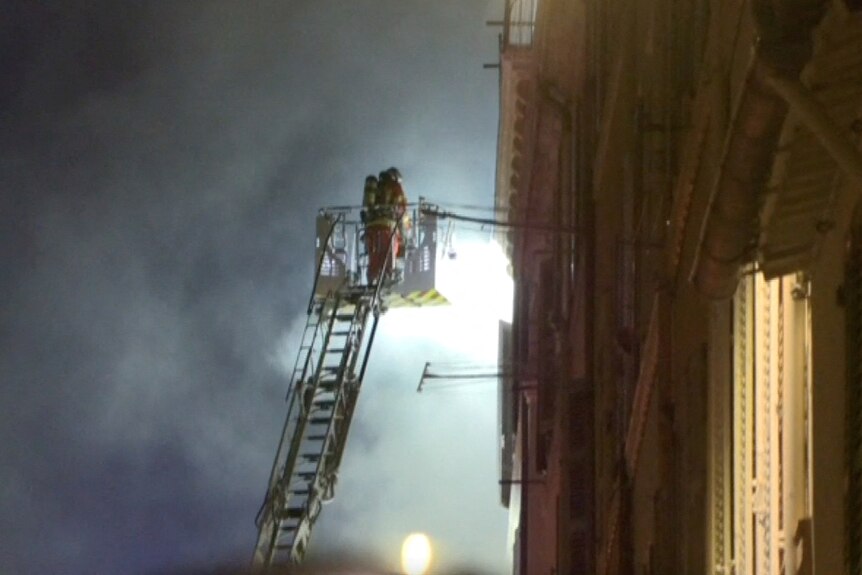 A firefighter stands on a ladder as they extinguish a building with smoke at night.