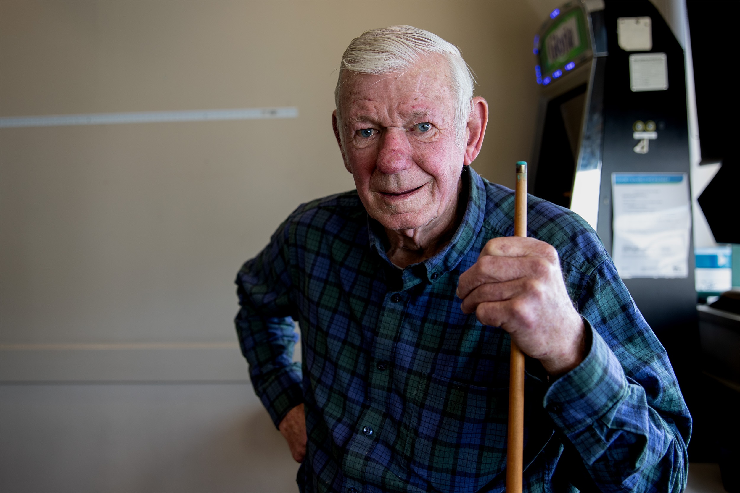 An old man with white hair looks ahead and smiles. He has one hand on his hip, another gripping a pool cue. He is smiling.