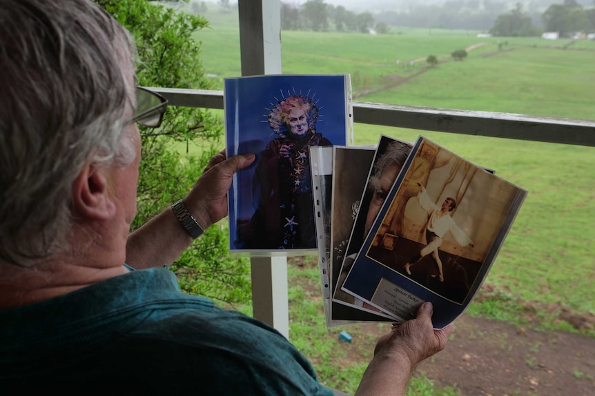 An older man holds old dancing photos in his hands, while sitting on a deck, with green fields in the distance.