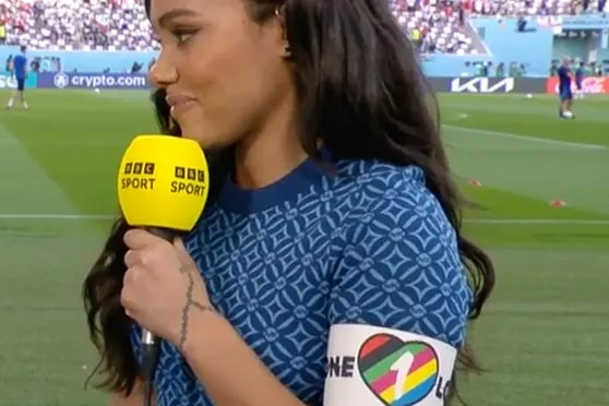 A news reporter stands on a soccer field at the World Cup and holds a yellow microphone while she conducts an interview