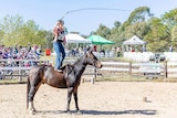 A teenager in jeans stands on a horse's bare back and cracks a whip as seated spectators look on.