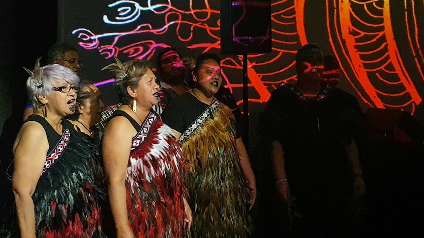 Women dressed in traditional feather dresses singing on stage wiht speaker and projected artwork of lines on black background