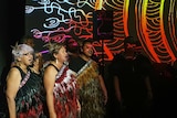 Women dressed in traditional feather dresses singing on stage wiht speaker and projected artwork of lines on black background