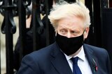 Boris Johnson's hair blows in the wind, he looks sideways, wearing a black face mask.