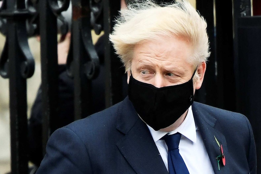 Boris Johnson's hair blows in the wind, he looks sideways, wearing a black face mask.