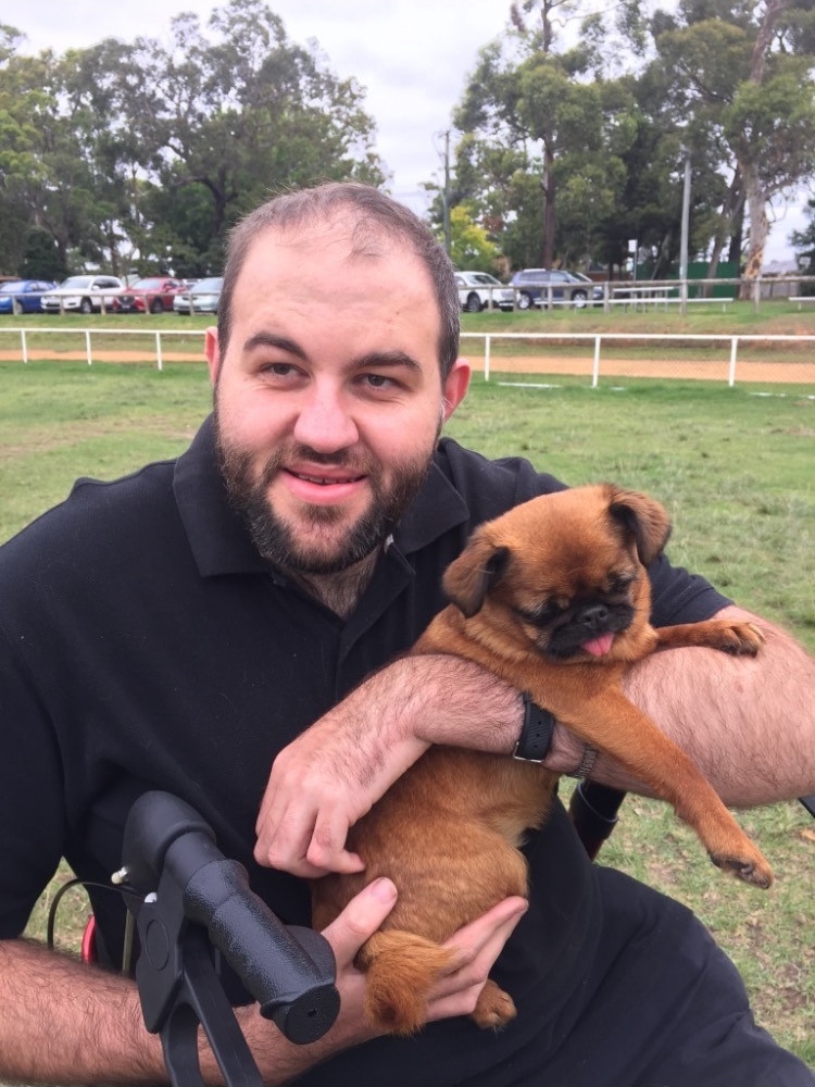 Lachlan, with sparse brown hair, a beard and a black t-shirt, sits in a park smiling and holding a small brown dog to his chest.