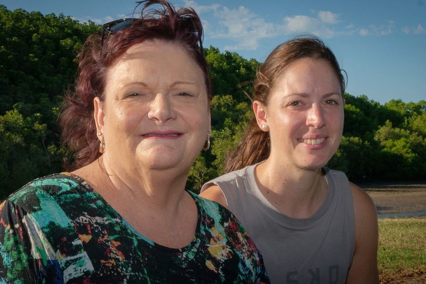 Two women sit together on a bench in front of lush green parkland. They are both smiling at the camera